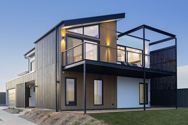 A modern two-storey residential home with a strongly articulated flat roof, dark vertical cladding, warm timber-look wall panels, expansive glazing, and a projecting upper-level balcony. Slim black steel posts and horizontal balcony rails create a lightweight contemporary facade, while a dark screened outdoor area extends along the right side. The house is photographed externally from the front, with a small landscaped verge and lawn visible.