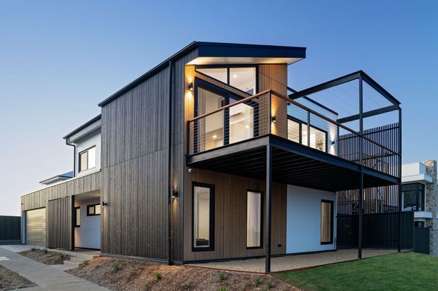 Exterior view of a contemporary multi-level residence with dark vertical cladding, crisp white wall sections, broad glazed openings, a projecting upper balcony, and a flat roof. The house includes a covered lower-level outdoor area, slim dark metal balustrades, and a compact landscaped frontage.