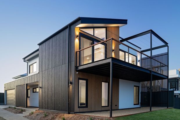 Contemporary two-storey residence with a dark vertical-clad facade, projecting upper-level balcony, extensive glazing, black metal balustrades, and a mix of flat and asymmetrical pitched roof forms. Warm interior lighting is visible through the windows at dusk.