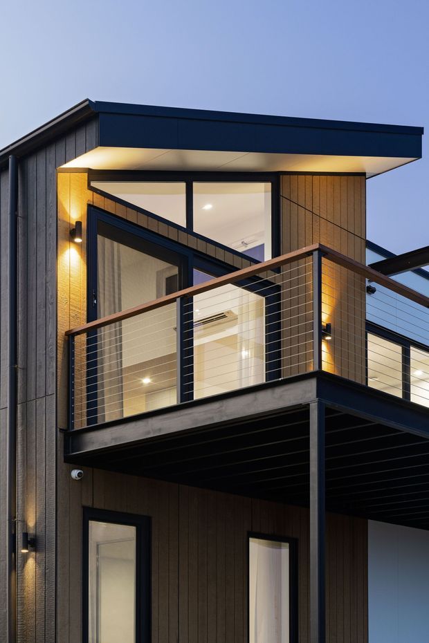Close-up dusk exterior view of a contemporary multi-storey residence with dark vertical cladding, a flat projecting roof, large glazed openings, and an elevated balcony with horizontal cable railing. Warm interior and exterior lighting contrasts with the dark blue-black facade and exposed balcony structure.