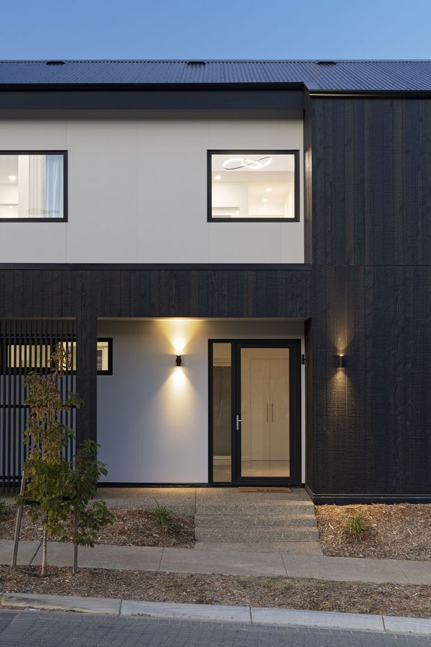 Street-facing view of a contemporary multi-storey townhouse facade at dusk. The building combines dark vertically textured cladding with light rendered wall panels, a dark metal pitched roof, black-framed windows, and a recessed entry with glazed double doors. Small landscaped garden beds and a concrete pathway lead to the entrance, with warm exterior wall lighting accenting the facade.