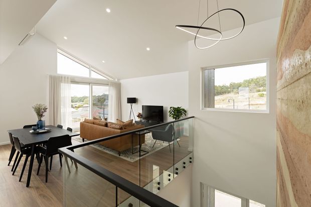 Bright open-plan upper-level living and dining area with a contemporary split-level layout. The room features light timber flooring, white walls and sloped ceilings, expansive glazing with sheer curtains, a black dining table, tan sofa, compact media area, glass balustrade overlooking a stairwell, and a textured warm-toned feature wall. A sculptural linear pendant adds a modern focal point.