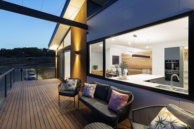 Contemporary outdoor deck adjoining a modern home, with timber decking, dark metal balustrades, lounge seating, and expansive black-framed glazing looking into a bright open-plan kitchen and living area. The home features dark cladding, warm timber wall detailing, a sharply pitched roofline, and integrated exterior lighting.