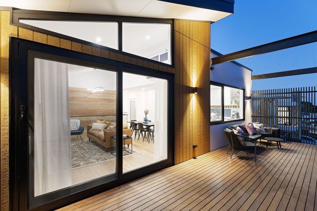 Contemporary rooftop or upper-level terrace with expansive timber decking, dark-framed sliding glass doors, warm vertical timber wall cladding, and a partially covered outdoor seating area. The interior living and dining spaces are visible through the glazing, while vertical privacy screening and a projecting steel-framed canopy define the terrace edge.