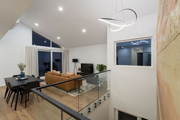 A bright contemporary open-plan living and dining area viewed from an internal stair landing. The space features a timber floor, black dining table and chairs, tan sofa, television, large glazed openings with curtains, a glass stair balustrade, and a sculptural linear pendant light. A textured stone or masonry feature wall is visible at right, with a lower level glimpsed below.