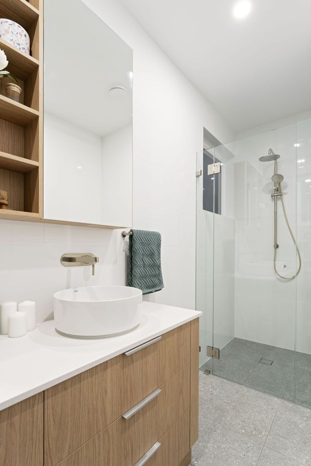 Bright contemporary bathroom featuring a wall-mounted timber vanity with white countertop and round vessel basin, mirrored cabinet, open timber shelving, wall-mounted tapware, and a frameless glass walk-in shower with brushed metal fixtures. Large light-grey floor tiles and white wall surfaces create a clean, spacious look.