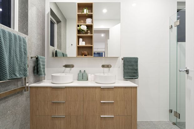 A bright, contemporary double-vanity bathroom with a timber-look floating vanity, two white vessel basins, wall-mounted tapware, a large mirrored cabinet with open timber shelving, and a frameless glass shower enclosure. Textured grey wall and floor finishes are paired with muted sage-green towels and accessories.