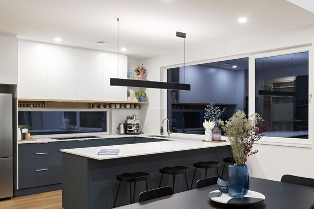 Contemporary open-plan kitchen and dining area with a large dark island, white engineered-looking benchtop, integrated cabinetry, pendant lighting, and dark-framed windows. The kitchen features dark navy-grey lower cabinetry, white walls and ceiling, timber flooring, open shelving, and a compact dining table in the foreground with decorative vases and dried flowers.