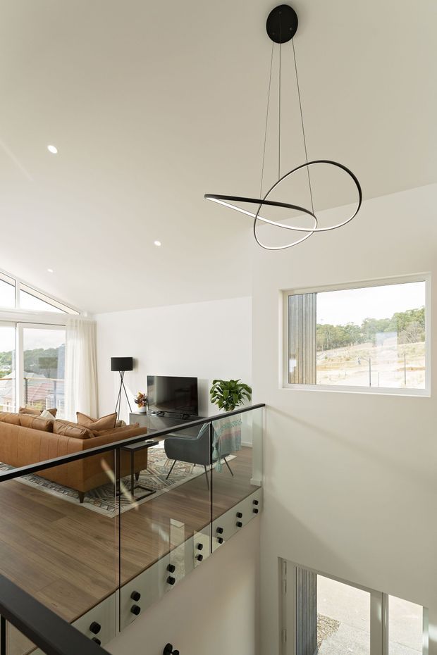 Bright contemporary open-plan living area viewed from a stair landing, featuring a double-height ceiling, large windows, glass stair balustrade, timber flooring, a tan sectional sofa, TV area, indoor plant, and a sculptural suspended loop pendant light.