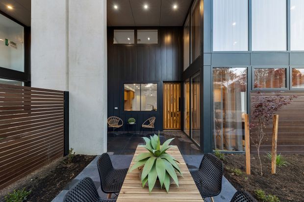 A contemporary residential entry courtyard viewed from outdoors, with a timber dining table and black woven chairs in the foreground, a large sculptural planter, dark vertical timber-clad walls, charcoal tiled paving, tall glazing, and a partially recessed front entrance. A pale concrete column and horizontal timber privacy screen frame the approach, while planted beds soften the architectural setting.