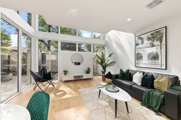 Bright contemporary living room with extensive floor-to-ceiling glazing and clerestory windows, opening to a leafy outdoor setting. The room features pale timber flooring, white walls and ceiling, a black leather sectional sofa with green, white and mustard cushions, a white oval coffee table, sculptural black lounge chairs, indoor plants, and a large framed architectural photograph. Natural light creates an airy, refined atmosphere.
