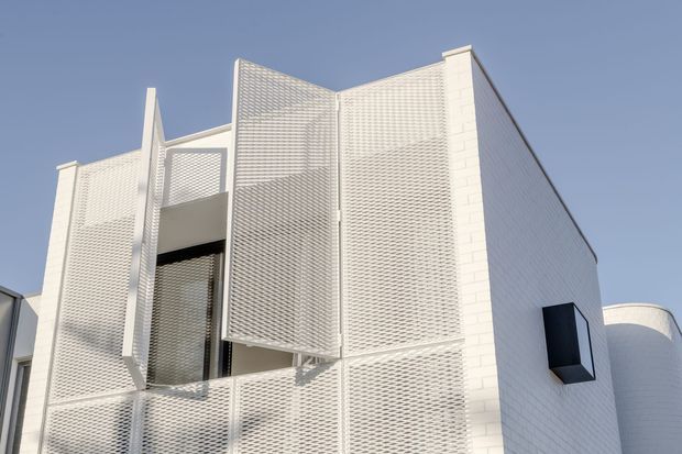 A contemporary white building exterior under a clear blue sky. The facade combines white brick walls with projecting perforated metal screens, deep recessed openings, and a dark projecting window box. A rounded building volume is partially visible at the right.
