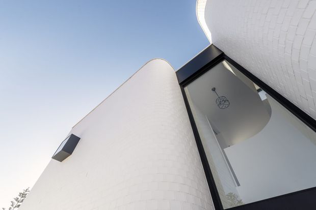 Upward-looking exterior architectural photograph of a contemporary building with curved white brick walls, sweeping rounded roof or parapet forms, and a tall black-framed glazed opening. The image emphasizes sculptural geometry, contrasting white masonry and dark metal framing against a pale sky.