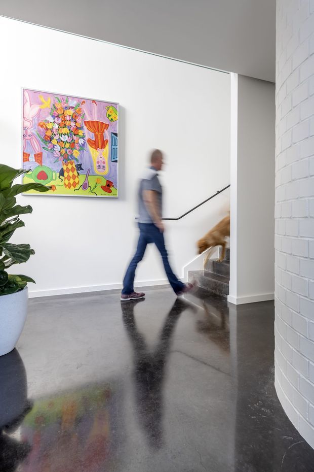 A bright contemporary interior hallway or landing with polished dark concrete flooring, white walls, and a curved white-painted brick wall. A stairway with a simple black handrail rises at the rear, while colourful artwork and a large potted plant add visual interest. Motion-blurred people are walking through the space.