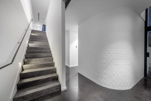 A clean, minimalist interior stairway with broad dark grey carpeted steps, a slim metal handrail, white walls, and a curved white-painted brick feature wall. The polished dark grey floor and concealed lighting create a contemporary, monochromatic atmosphere. A hallway or landing is visible beyond the stair, with a dark-framed opening at right.