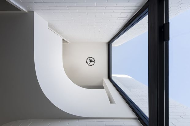 A dramatic upward-looking architectural view through a tall glazed opening, showing a curved white plaster wall and ceiling transition, white painted brickwork, a black-framed window or skylight, and a small round wall clock. The composition emphasises geometric lines, sculptural curves, and strong contrast between the white interior surfaces and dark glazing frame.