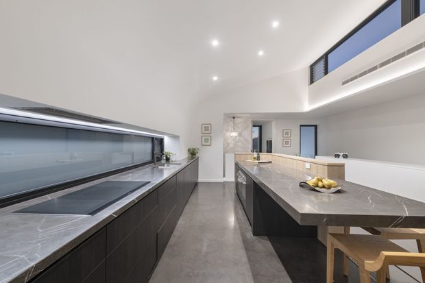 A sleek contemporary kitchen with a long central island and integrated timber breakfast seating. Dark flat-panel cabinetry contrasts with pale veined stone benchtops, a polished concrete floor, and extensive horizontal glazing along the rear wall. The room has a bright, minimalist fit-out with recessed lighting and a high-level clerestory window.