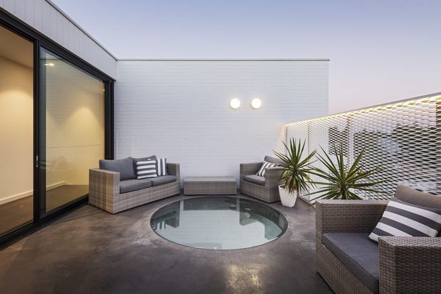 A contemporary rooftop-style outdoor terrace with polished concrete flooring, dark-framed floor-to-ceiling glazing, white horizontal wall cladding, and a perforated white privacy screen. Wicker lounge seating with grey cushions and striped pillows surrounds a circular glazed floor feature, with potted architectural plants adding greenery. The space has a refined, softly lit evening atmosphere.