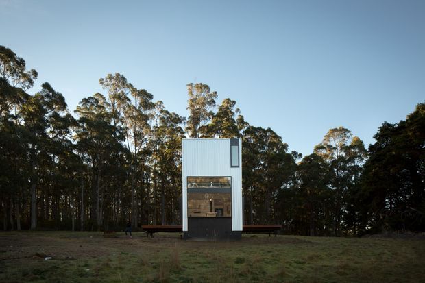 A compact, narrow modern pavilion set within a eucalyptus forest clearing. The simple rectangular form has pale corrugated cladding, a dark lower facade, a tall glazed opening revealing a small interior, and a narrow vertical window near the roofline. Timber benches or decks extend from both sides. The image is photographed from outdoors in soft natural light, with the forest and open grass surrounding the structure.