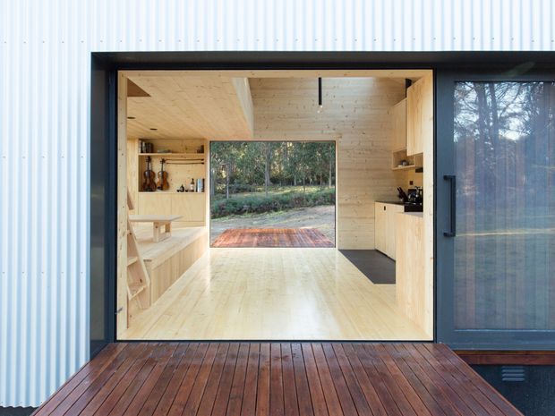 View from a dark timber deck through wide-open glazed doors into a compact, light-filled timber-lined interior. Pale timber flooring, walls, ceiling and built-in joinery frame a small kitchen and display/storage area, with a large opening beyond leading to a forested garden and a small outdoor deck. The exterior is clad in pale corrugated metal, with dark window and door framing.