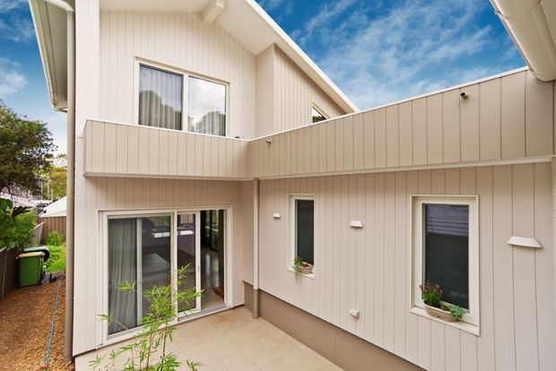 Exterior view of a contemporary multi-level residential building enclosing a small paved courtyard. The facade is clad in light beige vertical weatherboard-style panels with white-framed windows and doors, including large glazed openings at ground level. Gutters and downpipes are visible, along with a partly covered upper-level terrace or balcony edge. The image is brightly lit with a clean, neutral palette and landscaped planting at the courtyard perimeter.