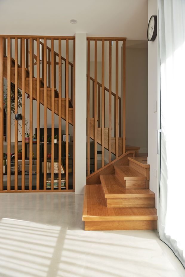 A bright interior stairway with warm timber stairs, matching timber balustrades and vertical slatted screening. The staircase rises behind a white structural column, with a shelving and living area visible through the screen. A large curtained window brings in strong natural light, creating striped shadows across the pale floor.