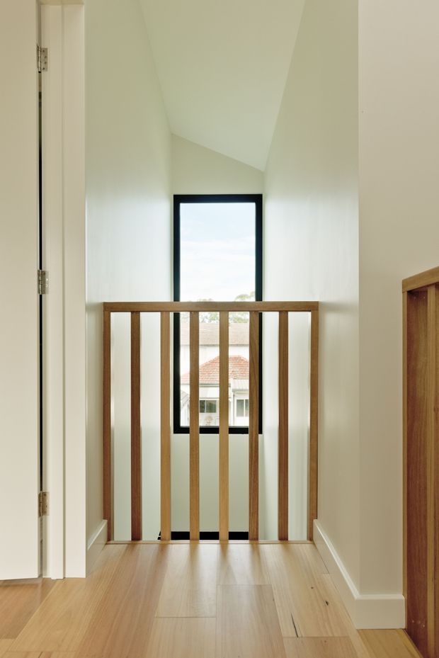 A bright residential upper-level landing or hallway overlooking a stairwell, with pale painted walls, warm timber flooring, a simple timber balustrade, and a tall black-framed window. A partial timber built-in cabinet is visible at right, while a tiled-roof neighbouring building can be seen through the window.