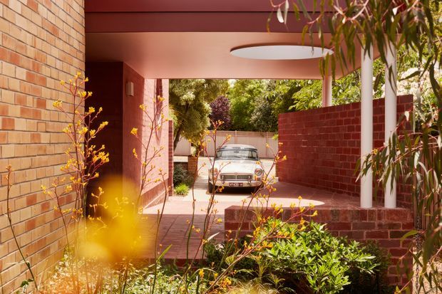 A warm, modern brick residential entry or carport viewed from a landscaped approach. Red and orange brick walls frame a covered passage with a deep red ceiling, a white cylindrical column, and a large oval recessed ceiling light. A vintage car is visible in the driveway beyond, while dense planting softens the foreground and edges. The composition emphasizes the sheltered connection between the street, garden, and home.