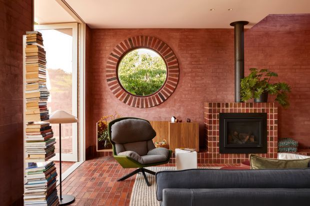 A warm, eclectic living room with exposed red brick walls, a matching brick-tiled floor, and a prominent freestanding wood-burning fireplace clad in small red and cream tiles. A circular window with radiating brick detailing forms a strong architectural feature wall. The room includes a dark upholstered sofa, sculptural lounge chair, timber sideboard, tall stack of books, indoor plant, and generous glazed opening, creating a cozy mid-century-inspired retreat.