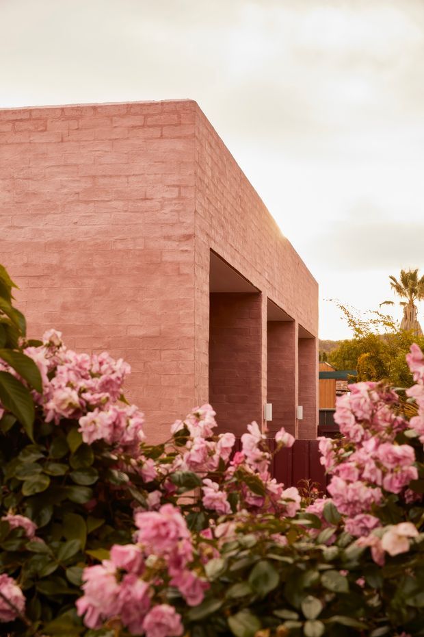 Exterior view of a modern, monolithic single-storey building with a flat roof and distinctive pink-toned exposed brick masonry. Deep recessed openings form a covered veranda or colonnade along the side, partly screened by abundant flowering garden vegetation. The warm, softly lit scene has a sculptural and minimalist character.