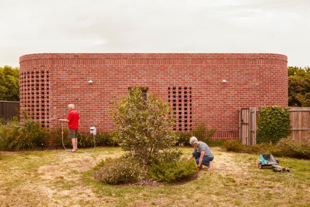 Exterior view of a distinctive single-storey curved red-brick building with rounded corners, patterned ventilation openings, and a simple flat roofline. The structure sits behind a landscaped garden with shrubs, timber fencing, and people gardening in the foreground.