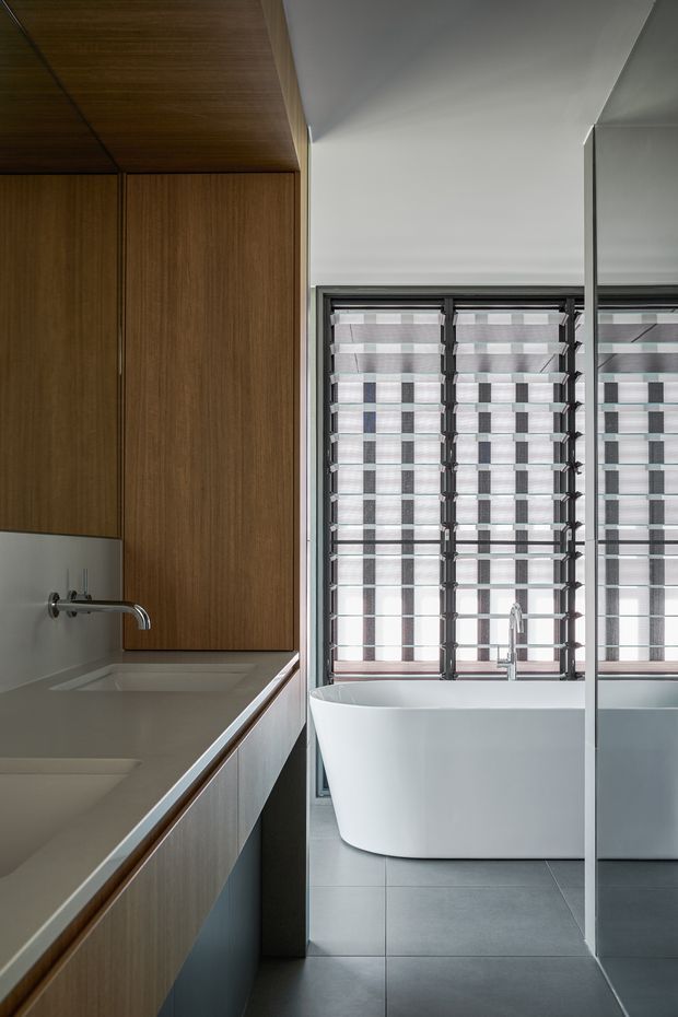 Contemporary bathroom interior featuring a freestanding white bathtub beside large louvred windows, a long timber-veneered vanity with integrated basins and wall-mounted chrome taps, pale walls, grey floor tiles, and a full-height mirror reflecting the bath and windows. The space is clean-lined, restrained, and naturally lit.