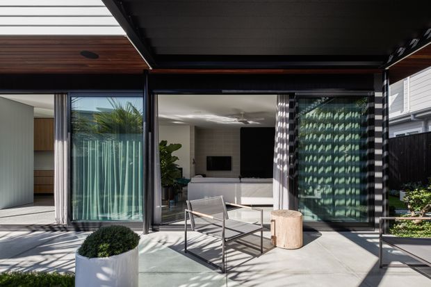 A contemporary covered outdoor living area opening directly into a bright interior living room. The space features large black-framed glazed sliding doors, a concrete patio floor, timber-lined ceiling sections, sheer curtains, a minimal outdoor lounge setting, and patterned sunlight filtering through screening or blinds. The interior includes a white built-in media console and television, with greenery visible around the patio.