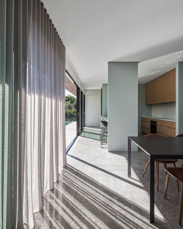 A bright, contemporary open-plan dining and kitchen interior viewed along full-height glazing. Sheer floor-to-ceiling curtains cast long shadows across a pale polished concrete floor. The kitchen features muted blue-grey vertical wall panelling, warm timber cabinetry, dark benchtops, and a compact island with stools. A dark dining table and timber chairs occupy the foreground, with a glimpse of outdoor greenery and a pool through the glazing.