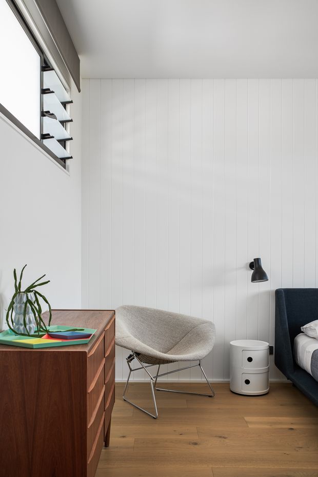 A bright, refined bedroom corner with white vertical wall panelling, warm timber flooring, and a mid-century-inspired furnishing arrangement. The scene includes a dark timber sideboard, sculptural upholstered lounge chair, black wall-mounted reading lamp, white cylindrical bedside table, and part of a dark upholstered bed. High-level louvred glazing brings in natural light.