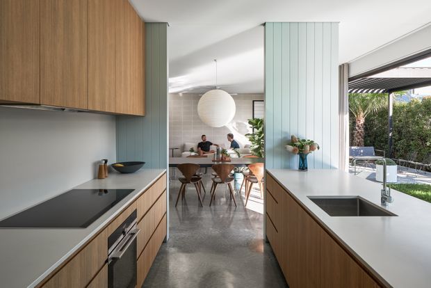 A bright, open-plan kitchen viewed along a central circulation aisle toward a dining area. The kitchen features warm vertical-grain timber cabinetry, pale blue-green fluted wall panels, white benchtops, an integrated black cooktop and oven, and a deep undermount sink. The dining space beyond has a timber table, sculptural pendant lights, and contemporary chairs. Large sliding glazing opens to a covered outdoor area and garden, creating a strong indoor-outdoor connection.