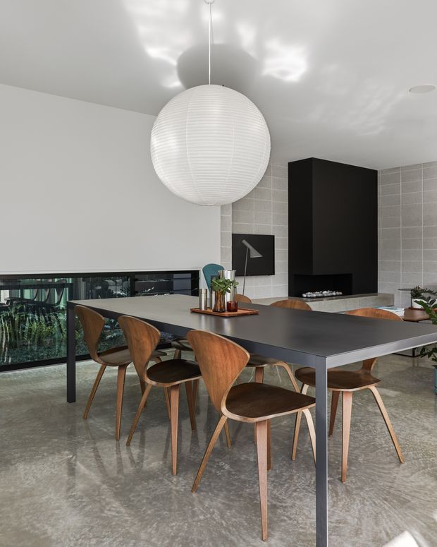 A contemporary dining area with a long dark dining table and curved wooden chairs, overlooking greenery through low horizontal glazing. The space features polished concrete flooring, white walls and ceiling, a large spherical paper pendant light, and a tiled feature wall with a tall black fireplace surround and built-in shelving. The restrained palette and reflective floor create a refined, modern atmosphere.