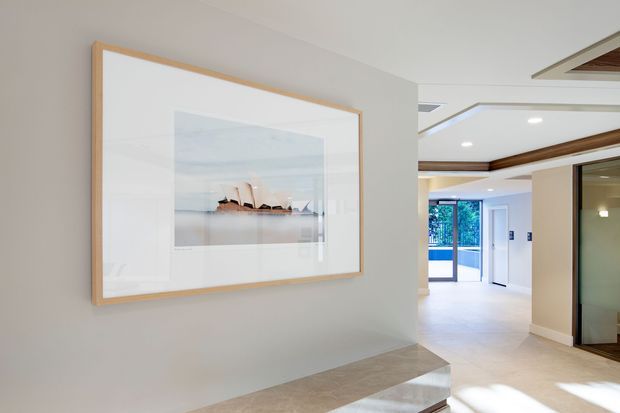 A bright contemporary interior hallway or entry area with pale neutral walls, a large framed artwork, light stone or tile flooring, recessed ceiling lights, timber ceiling beams, and a glazed doorway leading toward an outdoor or adjacent area. Built-in cabinetry or panelled doors are visible along the right side.
