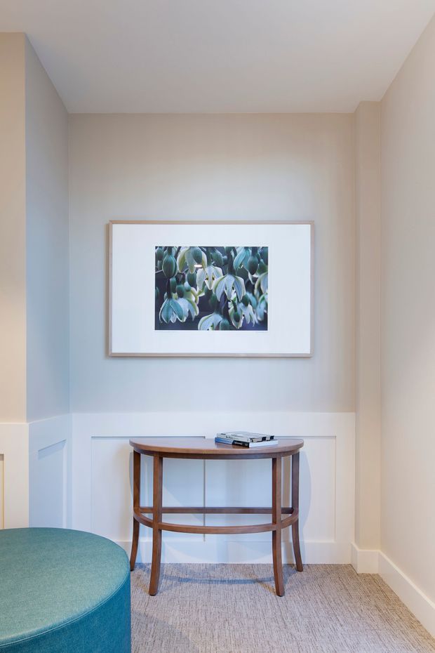 A refined residential hallway or entry nook with warm off-white wall panelling, a framed botanical artwork, and a slim curved timber console table with a lower shelf. A teal upholstered rounded seat is partially visible in the foreground, set over a neutral textured carpet. The space is softly lit and understated.