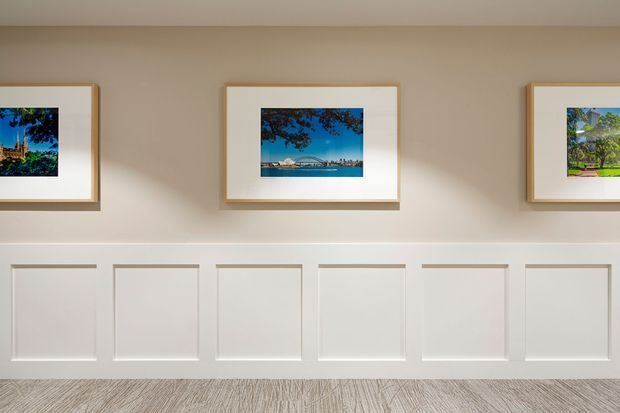 A softly lit interior wall featuring evenly spaced framed landscape photographs above a series of white rectangular wall panels. The lower floor area has a light, textured finish, creating a clean and understated gallery-like corridor or reception setting.