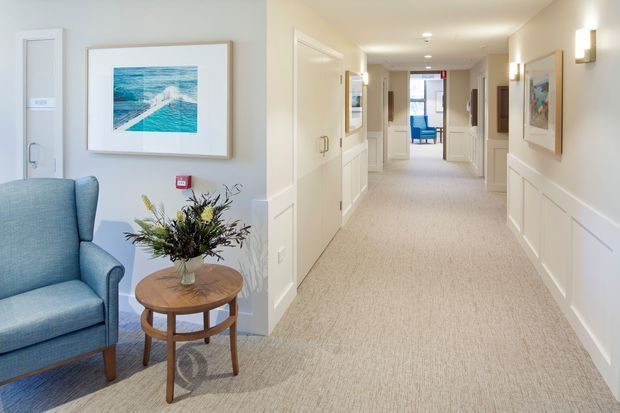 A bright, refined interior showing a long carpeted hallway with white painted wall panelling, framed artwork, wall sconces, and multiple doorways. In the foreground is a small sitting area with a blue upholstered armchair, round timber side table, and floral arrangement. A blue chair and glazed doorway are visible at the far end.