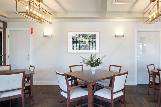 A refined dining room with a large rectangular dark timber dining table surrounded by upholstered timber-framed chairs. The room has pale walls, herringbone timber flooring, framed artwork, wall sconces, and prominent brass-and-glass geometric pendant lights. Additional dining seating is visible at the edges, creating a polished hospitality-style interior.