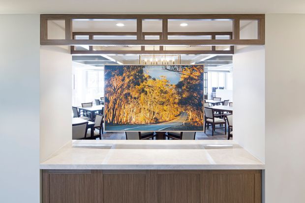 A contemporary built-in media and display wall viewed from a bright interior. The central large screen shows an autumn forest road, flanked by glimpses of dining or lounge seating behind it. A pale stone or engineered-stone ledge sits above dark vertically grained timber cabinetry, with matching timber-framed clerestory-style glazing and overhead architectural framing. Recessed lighting and a linear chandelier add illumination.