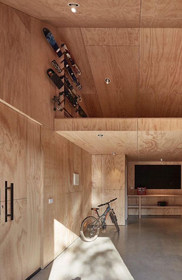 A compact garage or gear-storage space fully lined in warm plywood, with integrated shelving and a wall-mounted television at the rear. Skis are displayed high on the wall, while a bicycle occupies the polished concrete floor. Recessed spotlights, flush plywood cabinetry, and a large black vertical door handle contribute to a clean, highly integrated design.