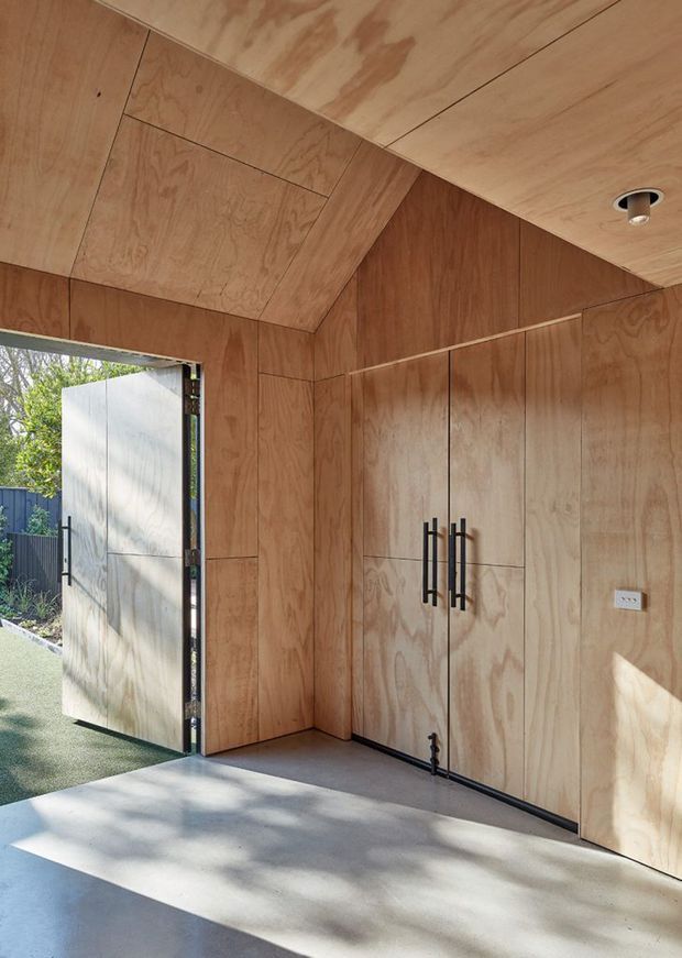 A compact timber-lined interior, likely an entry or utility space, with plywood wall and ceiling panelling, integrated full-height storage cabinetry, black vertical handles, a concrete floor, recessed ceiling lighting, and an open exterior door leading to a garden or yard.