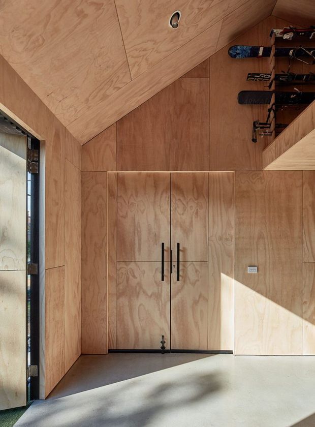 A compact, plywood-lined interior with full-height built-in cabinetry and double doors, viewed at an angle. Warm timber wall and ceiling panels create a cohesive enclosure, while a wall-mounted rack displays skis or similar sporting equipment. A narrow glazed opening brings in strong natural light, contrasting with the dark metal door handles and grey floor.