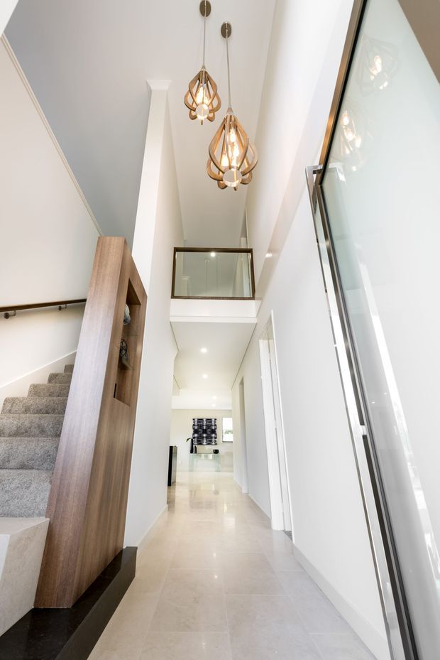 Bright, contemporary multi-level hallway with a tall stairwell and double-height void. The space features pale polished floor tiles, white walls, a carpeted staircase with dark handrail, integrated mid-tone timber shelving beside the stairs, glass balustrading, and two sculptural pendant lights. A mezzanine or upper-level opening is visible above the corridor.