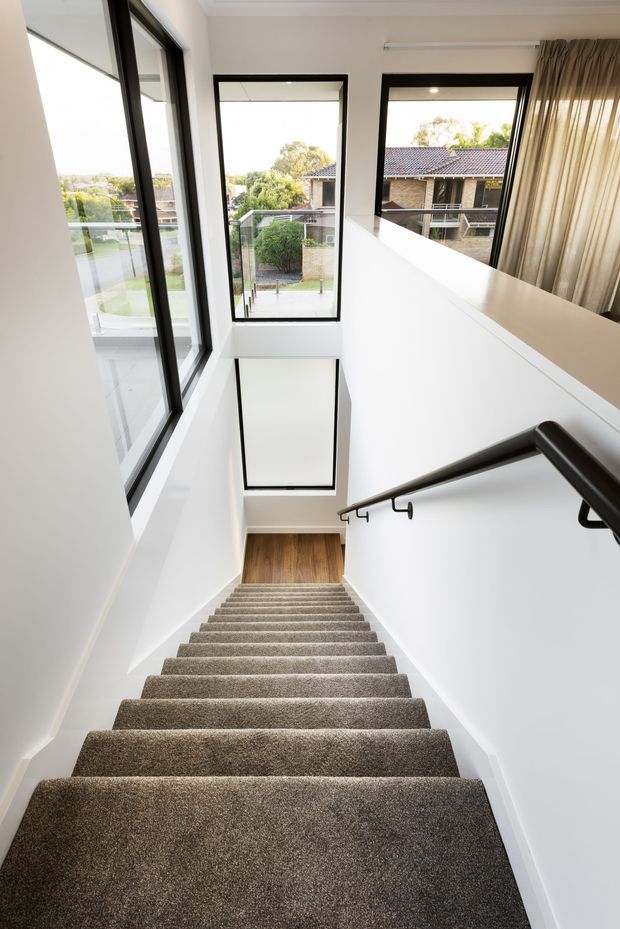 Bright, narrow residential stairway viewed from the upper level, with a long flight of carpeted stairs descending to a timber-finished landing. White walls and ceiling are paired with large black-framed windows, a glass balustrade, and a dark handrail, creating strong natural light and views to neighbouring homes.