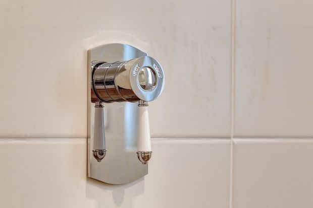 Close-up of a polished chrome wall-mounted bathroom fixture with two hanging pull handles, installed on a glossy white tiled wall. The fixture has a rounded, retro-inspired form and a reflective metal finish.