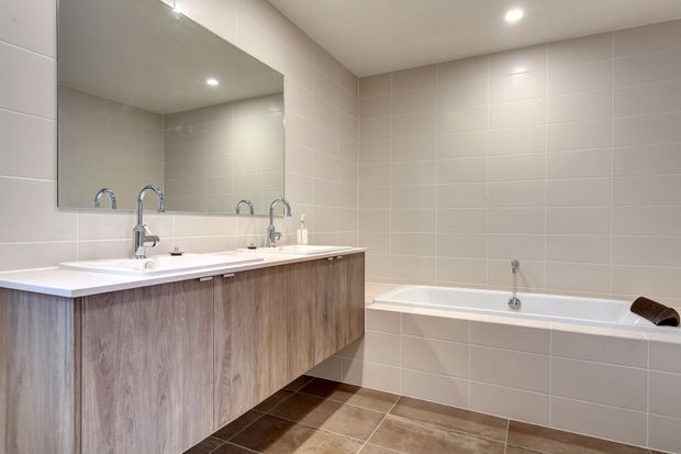 A bright, contemporary bathroom with a long double vanity, wood-look fixed cabinetry, white benchtop, wall-to-wall mirror, tiled walls, tiled floor, and a built-in bathtub. Recessed ceiling lighting provides even illumination.